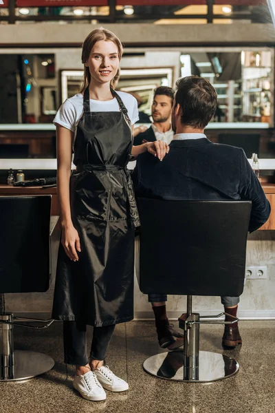 Schöne junge Friseurin lächelt in die Kamera, während männliche Klientin im Stuhl im Schönheitssalon sitzt — Stockfoto