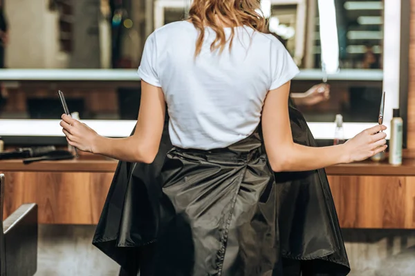 Vista posterior de peluquería sosteniendo peine y tijeras, mientras que el cliente masculino sentado en la silla en el salón de belleza - foto de stock