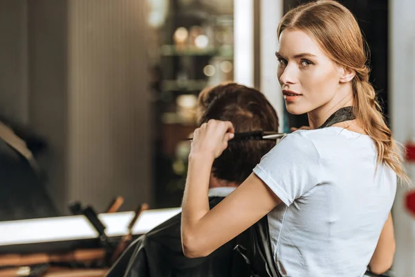 Bellissimo giovane parrucchiere guardando la fotocamera mentre pettina e taglia i capelli al cliente maschile nel salone di bellezza — Foto stock