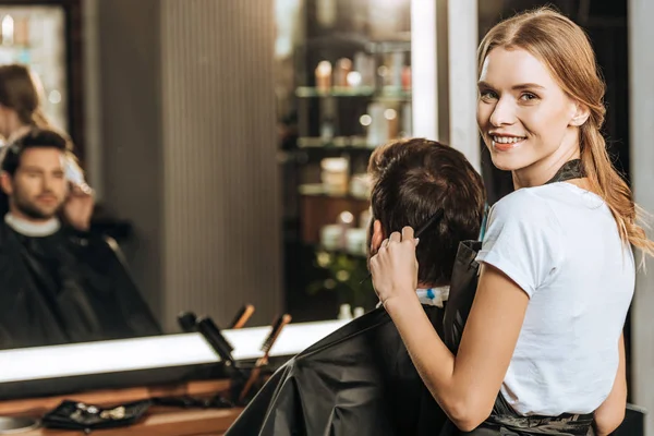 Schöne junge Friseurin lächelt in die Kamera, während sie dem männlichen Kunden im Schönheitssalon die Haare kämmt — Stockfoto