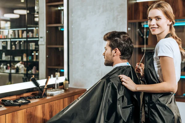 Friseur lächelt in die Kamera, während er einem hübschen jungen Mann im Schönheitssalon die Haare schneidet — Stockfoto