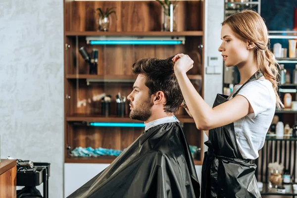 Vue latérale de coiffeur couper les cheveux à beau jeune homme dans le salon de beauté — Photo de stock
