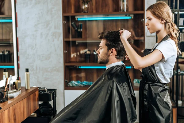 Vue de côté du coiffeur couper les cheveux à beau jeune homme dans le salon de beauté — Photo de stock