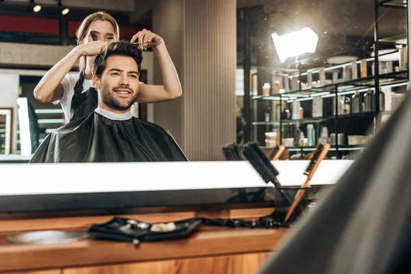 Отражение в зеркале молодого парикмахера, стригущего волосы красивому улыбающемуся мужчине в салоне красоты — стоковое фото