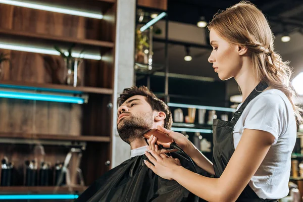 Молодий перукар обрізає бороду красивому клієнту в салоні краси — стокове фото