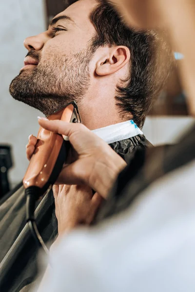 Close-up partial view of hairdresser using electric hair trimmer and cutting beard to client in beauty salon — Stock Photo