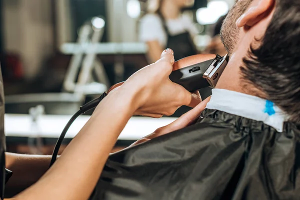 Recortado tiro de peluquería utilizando cortador de pelo eléctrico y corte de barba al cliente en salón de belleza - foto de stock
