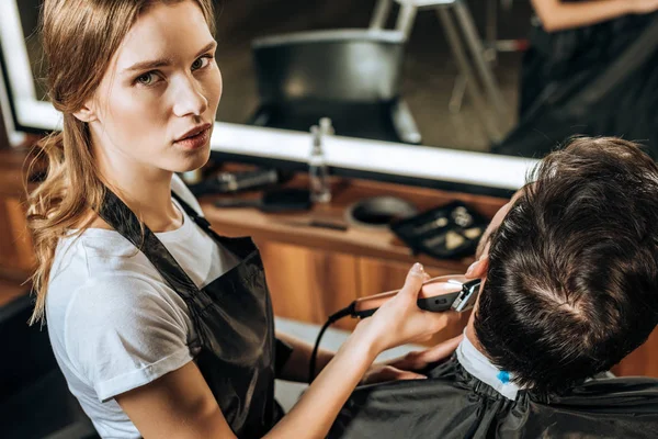 Vista de ángulo alto de la peluquería joven usando cortador de pelo eléctrico y mirando a la cámara mientras trabaja con el cliente en el salón de belleza - foto de stock