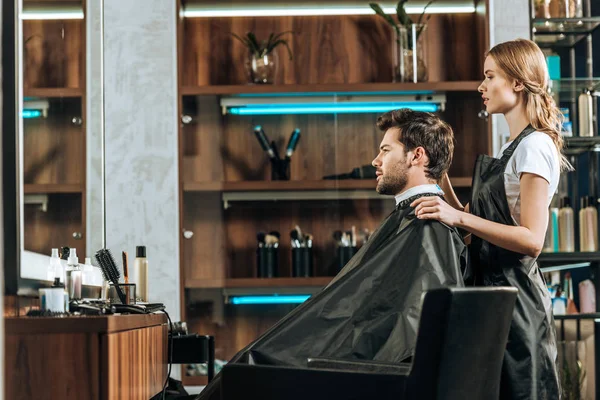 Vue latérale du coiffeur féminin et beau jeune homme regardant miroir dans le salon de beauté — Photo de stock
