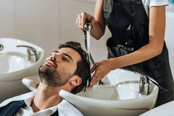 Recortado tiro de peluquero lavando el pelo a guapo joven en salón de belleza - foto de stock