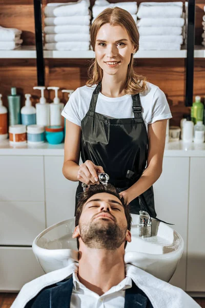 Hermosa peluquera sonriendo a la cámara mientras lava el cabello a un hombre guapo en el salón de belleza - foto de stock