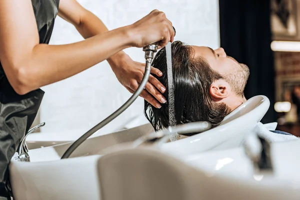 Teilansicht des Friseurs, der einem hübschen jungen Mann im Schönheitssalon die Haare wäscht — Stockfoto