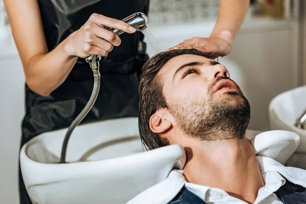 Vista parcial de peluquero lavado de cabello a guapo joven cliente masculino en salón de belleza - foto de stock