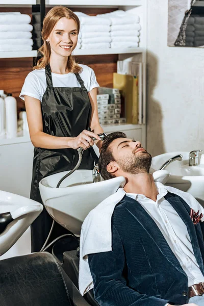 Joven peluquero sonriendo a la cámara mientras se lava el cabello a guapo joven cliente masculino en salón de belleza - foto de stock
