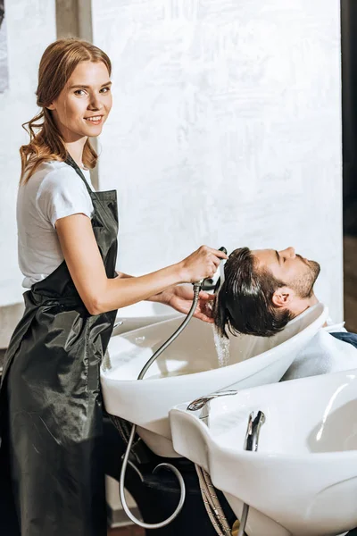 Cabeleireiro sorrindo para a câmera enquanto lava o cabelo para bonito jovem cliente masculino no salão de beleza — Fotografia de Stock