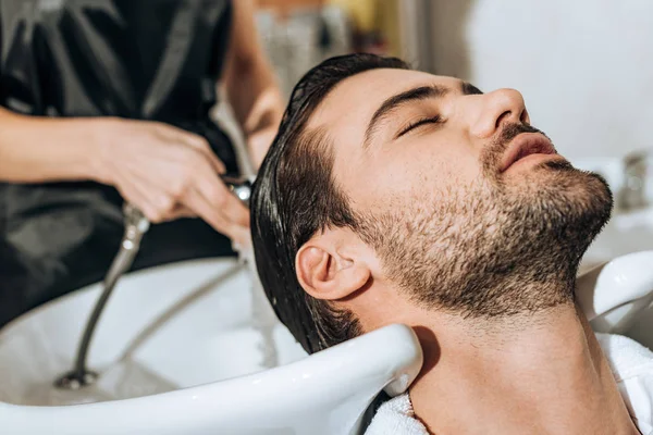 Primer plano vista parcial de peluquero lavando el cabello a guapo joven cliente masculino en salón de belleza - foto de stock