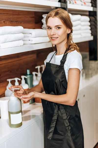 Hermosa joven estilista sosteniendo loción y sonriendo a la cámara en el salón de belleza - foto de stock