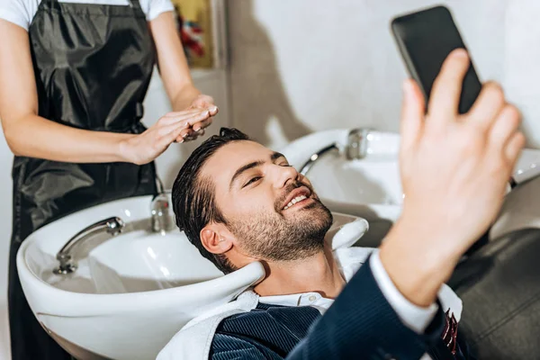 Обрезанный снимок парикмахера, моющего волосы улыбающемуся мужчине, делающему селфи со смартфоном в салоне красоты — стоковое фото