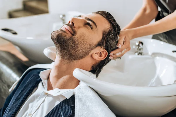 Vista parziale del parrucchiere che lava i capelli al bell'uomo sorridente nel salone di bellezza — Foto stock
