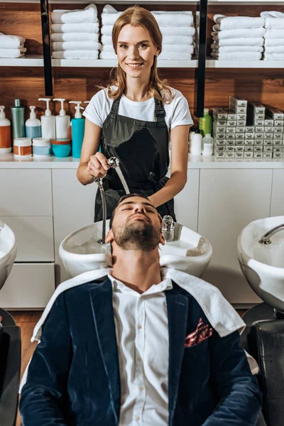 Bella giovane parrucchiere sorridente alla macchina fotografica mentre lavava i capelli a bell'uomo nel salone di bellezza — Foto stock