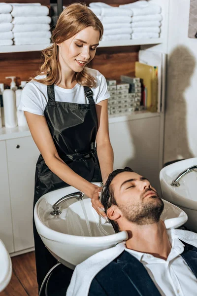 Lächelnde Friseurin, die einem hübschen jungen Mann im Schönheitssalon die Haare wäscht — Stockfoto