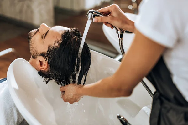 Vista parcial del estilista lavando el cabello a un joven guapo en el salón de belleza — Stock Photo