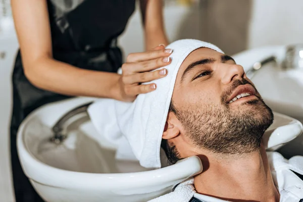 Partial view of hairdresser washing hair to smiling handsome man in beauty salon — Stock Photo