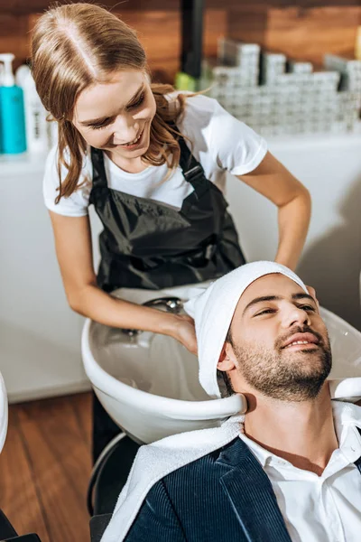 Visão de alto ângulo de cabelo jovem cabeleireiro lavar o cabelo para homem bonito sorridente no salão de beleza — Fotografia de Stock