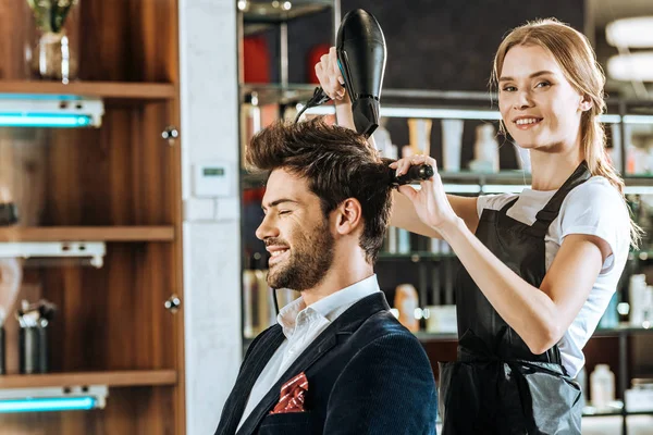 Hermoso joven peluquero sonriendo a la cámara mientras se seca y peina el cabello a un cliente guapo en el salón de belleza - foto de stock