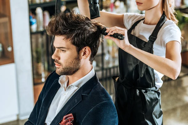 Recortado disparo de peluquería joven peinando y secando el cabello a guapo cliente en salón de belleza - foto de stock