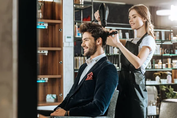 Beautiful young hairdresser drying hair to handsome smiling man in beauty salon — Stock Photo