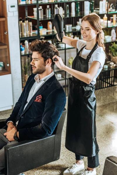 Visão de alto ângulo do cabelo de secagem de cabeleireiro feminino sorridente para jovem bonito no salão de beleza — Fotografia de Stock