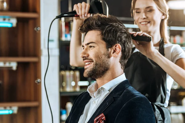Sonriente joven peluquero secando el pelo a guapo feliz hombre en salón de belleza - foto de stock