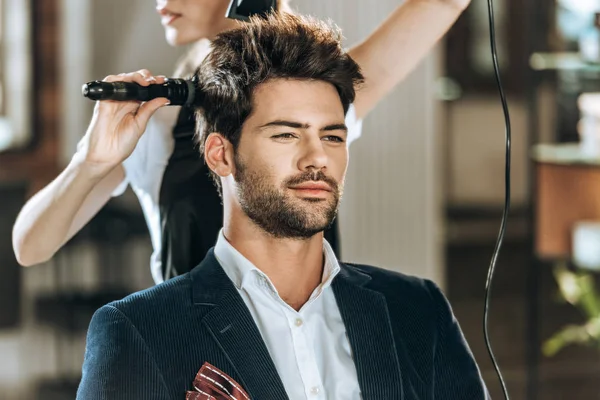 Recortado tiro de peluquero peinando y secando el cabello a guapo joven en salón de belleza - foto de stock