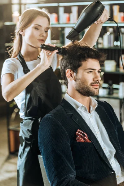 Joven peluquero peinando y secando el cabello al cliente guapo en salón de belleza - foto de stock