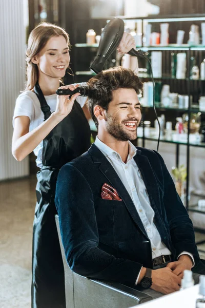Young hairstylist combing and drying hair to smiling handsome client in beauty salon — Stock Photo