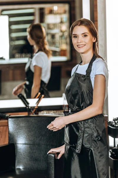 Attractive young hairstylist showing empty chair and smiling at camera in beauty salon — Stock Photo
