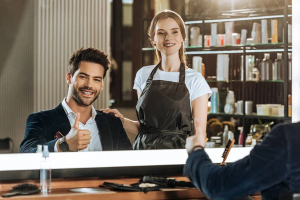 Beautiful hairdresser standing near handsome smiling client showing thumb up in beauty salon — Stock Photo
