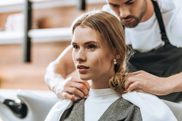 Hairstylist putting towel on client shoulders and young woman looking away in beauty salon — Stock Photo