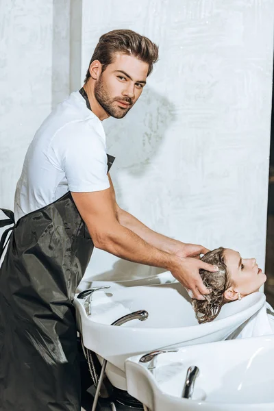 Handsome young hairstylist looking at camera and washing hair to attractive woman in beauty salon — Stock Photo