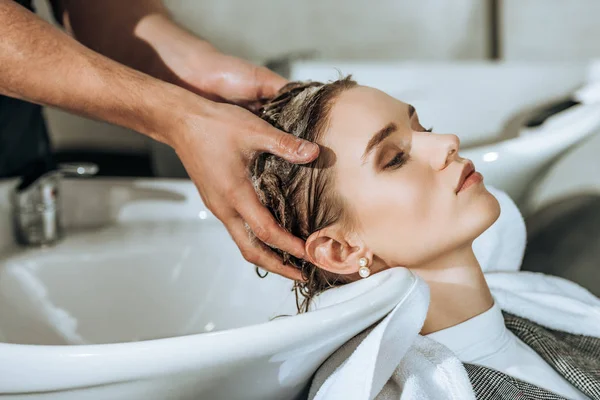 Vue partielle de coiffeur laver les cheveux à la jeune femme attrayante dans le salon de beauté — Photo de stock
