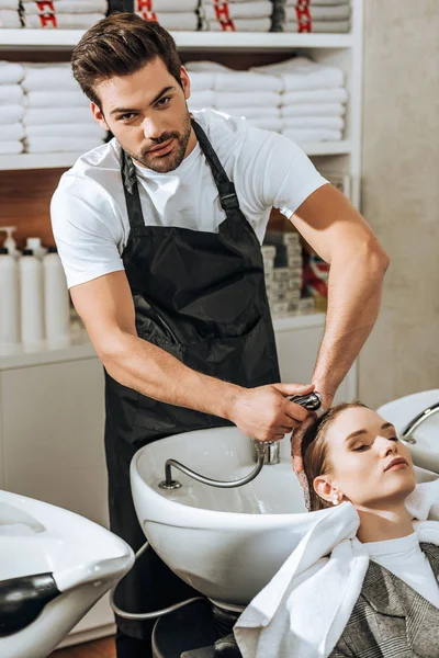 Bonito jovem cabeleireiro olhando para a câmera enquanto lavar o cabelo para mulher no salão de beleza — Fotografia de Stock