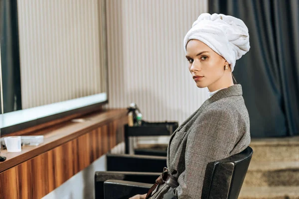Attraente giovane donna con asciugamano sulla testa seduta e guardando la fotocamera nel salone di bellezza — Foto stock