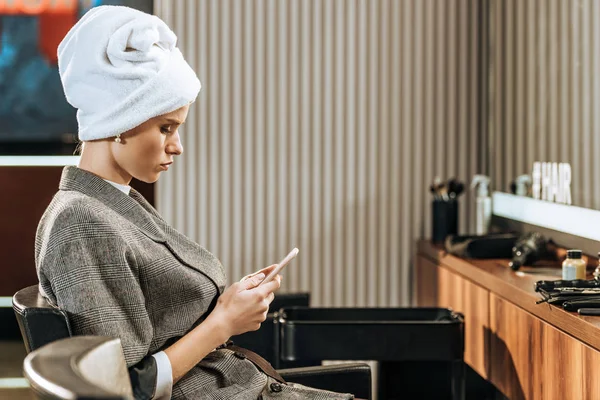 Приваблива молода жінка з рушником на голові сидить і використовує смартфон в салоні краси — стокове фото
