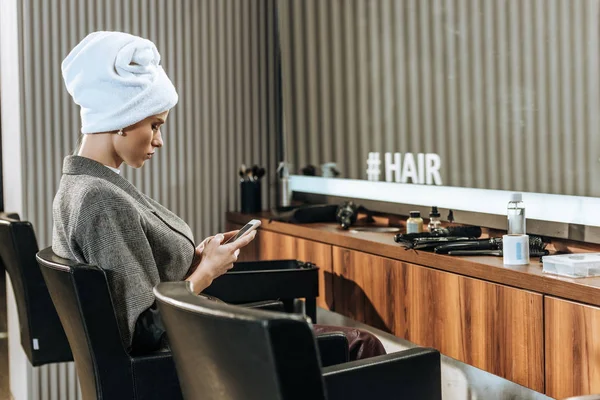 Красива молода жінка з рушником на голові сидить і використовує смартфон в салоні краси — стокове фото