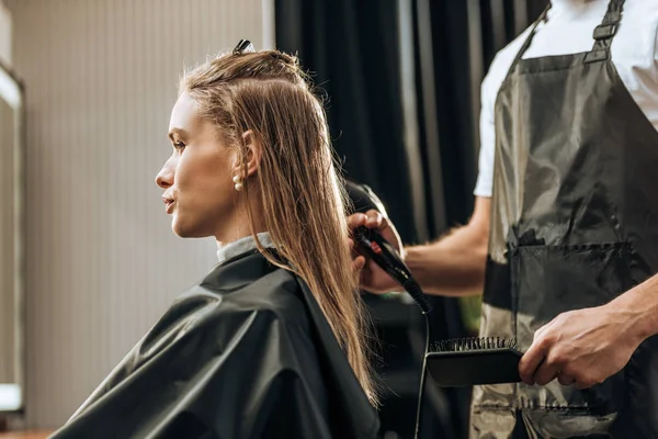 Обрізаний знімок перукаря, який тримає щітку для волосся та сушіння волосся привабливій дівчині в салоні краси — стокове фото
