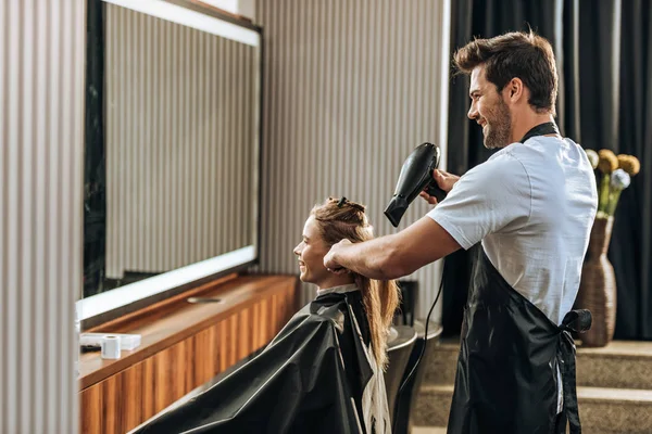 Visão lateral de sorrir jovem cabeleireiro secagem cabelo para menina feliz no salão de beleza — Fotografia de Stock