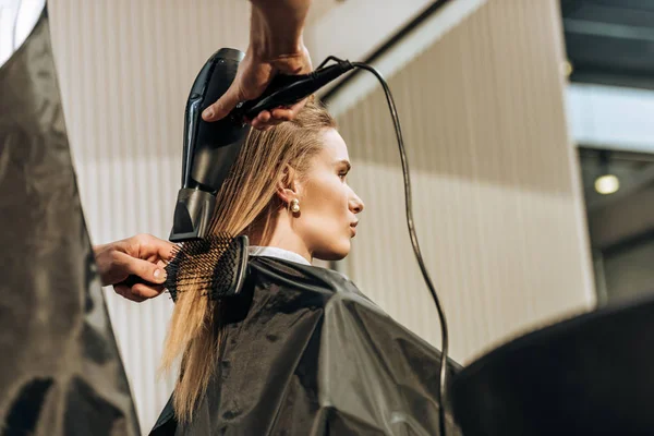 Vue à angle bas de coiffeur séchage des cheveux à la fille attrayante dans le salon de beauté — Photo de stock