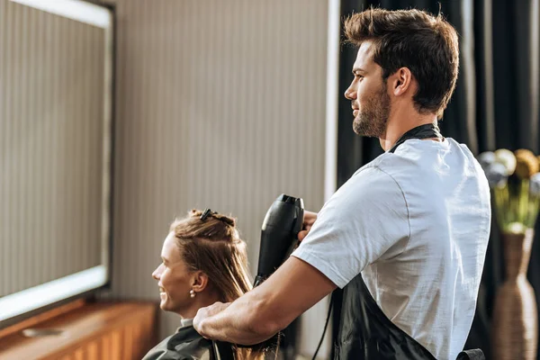 Красивый молодой парикмахер сушит волосы привлекательной девушке в салоне красоты — стоковое фото