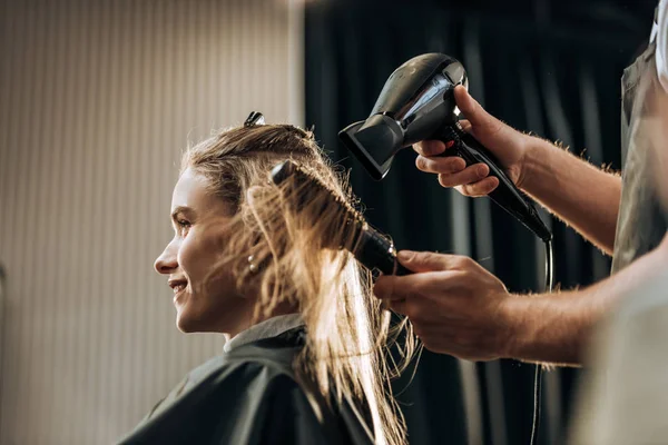 Plan recadré de coiffeur séchant les cheveux à la fille souriante dans le salon de beauté — Photo de stock